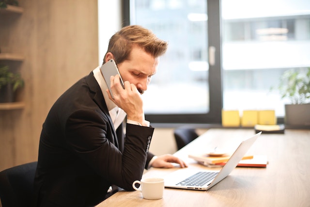 A landlord in a black suit who is receiving an emergency maintenance call from a tenant sits at their desk and arranges repair services on their laptop